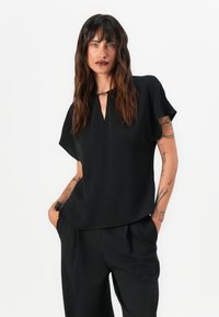 Blusa nera a maniche corte con scollo a V e dettaglio a chiave frontale, realizzata in tessuto morbido, abbinata a pantaloni neri larghi.