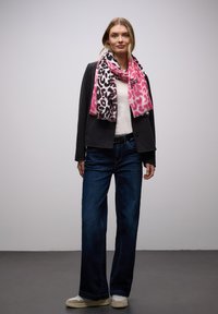 Femme portant une veste noire, une écharpe imprimé léopard rose et noir, un haut blanc, un jean bleu et des baskets blanches, debout devant un fond uni.