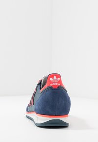 Granatowy zamszowy sneaker z różowymi akcentami. Posiada teksturowane tylne wstawki, trzy czerwone paski oraz biały środek podeszwy z gumową podeszwą.