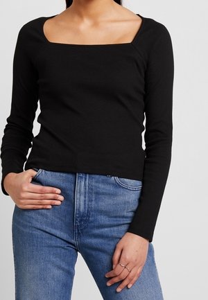 Kvinde iført en sort langærmet top med firkantet halsudskæring og blå højttaljede jeans, med den ene hånd i lommen og den anden hvilende på låret.