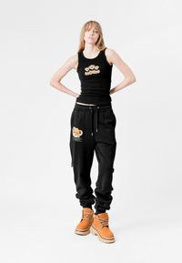Top negro sin mangas con un diseño de oso, combinado con pantalones de chándal negros que tienen un parche de oso de peluche y botas amarillo-naranjas. Textura suave en todo.