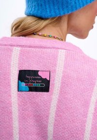 Pruhovaný růžový svetr s texturovaným pletením, na kterém je černá nálepka s nápisem "štěstí v pokroku" v barevném písmu.