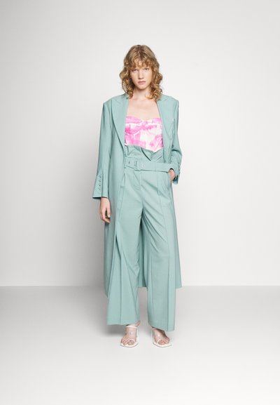 Um fato em verde menta, ajustado, com calças de perna larga, apresentando uma cintura com cinto e um casaco longo. Por baixo, há uma blusa em tie-dye rosa e branca. Sandálias transparentes completam o visual.