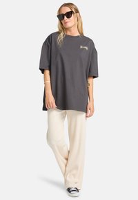 Overdimensioneret grå t-shirt med et logo, parret med cremefarvede vidde bukser og sorte sneakers. Akkompagneret af sorte solbriller.