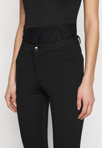 Svarta figurnära byxor med en slät textur, framfickor, dragkedja samt en metallknapp för knäppning vid midjan.
