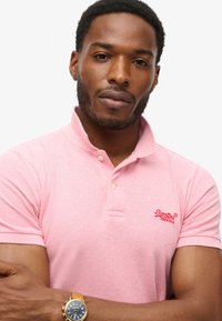 Różowa koszulka polo z krótkimi rękawami, prążkowanym kołnierzykiem oraz wyhaftowanym czerwonym logo. Model ma zegarek z brązowym paskiem.