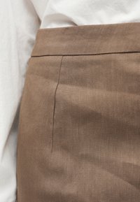 Gros plan d'une personne portant une jupe en lin marron clair taille haute et une chemise blanche avec la ceinture de la jupe visible.