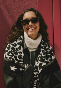 Sort læderjakke, cremefarvet turtleneck og et strikket sort og hvidt leopardmønsteret sjal med stjernedetaljer. Modellen bærer mørke solbriller.
