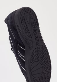 Čierna atletická obuv s textúrovanou gumovou podrážkou. Obsahuje biele prúžky a sieťovinový zvršok. Branding viditeľný na povrchu podrážky.