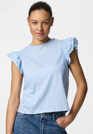 Fiatal nő, hátrafésült hajjal, világoskék rövid ujjú fodros szemenyílásos blúzt és kék farmernadrágot visel, keze zsebbe dugva.