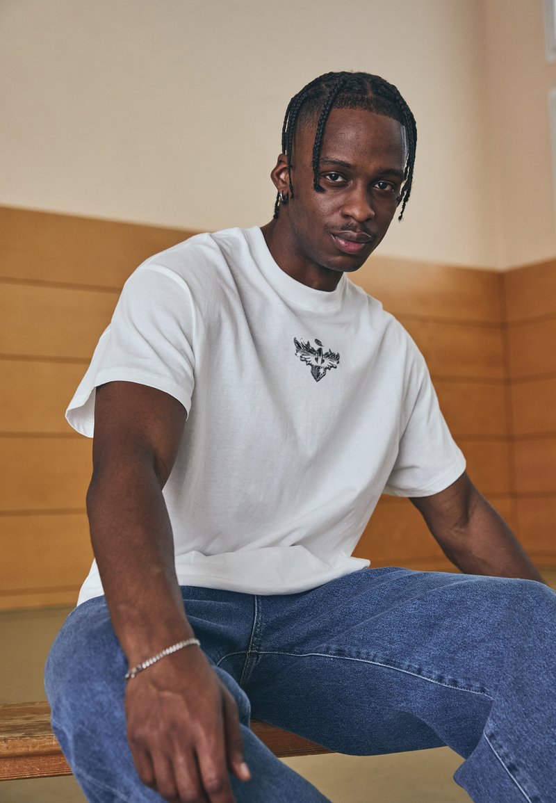 Hvid bomulds t-shirt med et sort grafisk design på brystet. Parret med blå denim jeans og siddende på en træbænk i en træningssal.