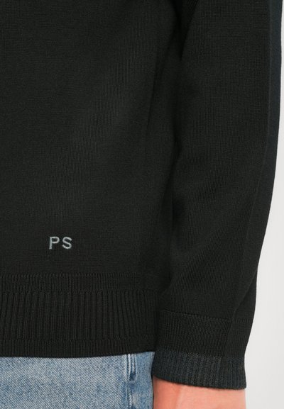Schwarzer Strickpullover mit Ärmel und unterem Saum, auf dem in der Nähe des Saums "PS" gestickt ist, kombiniert mit hellblauen Denim-Jeans.