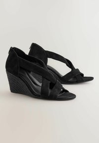 Sandales compensées en suédine noire avec des brides entrecroisées. Elles présentent un talon compensé tissé noir texturé et un design à bout ouvert. Fermeture éclair au niveau du talon.