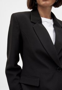 Melnā žakete, kas izgatavota no teksturēta auduma, ar iekļautu apmali, vienu pogu aizdari un priekšējo kabatu. Uzvilkta virs baltā topa.