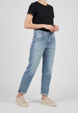 Femme portant un jean bleu clair retroussé, un t-shirt noir à manches courtes et des baskets beige, debout avec les mains jointes près de la taille.
