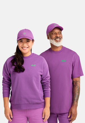 Lachende man met witte baard en vrouw met bijpassende paarse petten, tops en broeken met kleine groene logo's op de borst en pet.