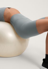 Höga shorts i midjehöjd av mjukt grått tyg, i knä-längd. Slät textur med plana sömmar, burna medan personen sitter på en ljus färgad gymboll.