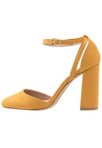 Żółty zamszowy but na wysokim obcasie z długim noskiem i paskiem na kostce z klamrą. Projekt z blokowym obcasem zapewnia stabilność i komfort.
