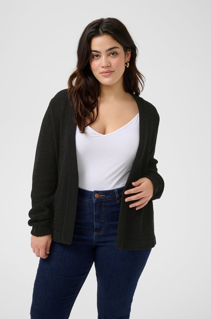 Cardigan en tricot vert foncé, à l'avant ouvert, en tissu texturé, à manches longues. Associé avec un top blanc à col en V et un jean taille haute bleu.