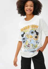 Hvid bomulds T-shirt med et farverigt grafisk design med blomster, svampe og teksten "GROOVY VIBES" i fede orange og blå bogstaver.