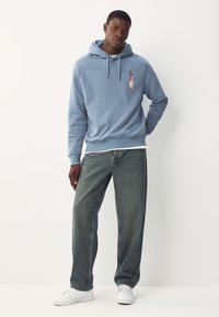 Mand iført lyseblå hættetrøje med grafik af en flaske stærk sauce, løse, falmede sorte jeans og hvide sneakers, stående op ad en hvid væg.