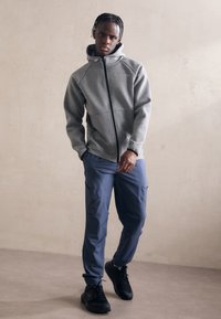 Junger Mann mit geflochtenem Haar, der einen grauen Zip-Hoodie, blaue Cargohosen und schwarze Sneakers trägt und vor einer schlichten beigen Wand steht.