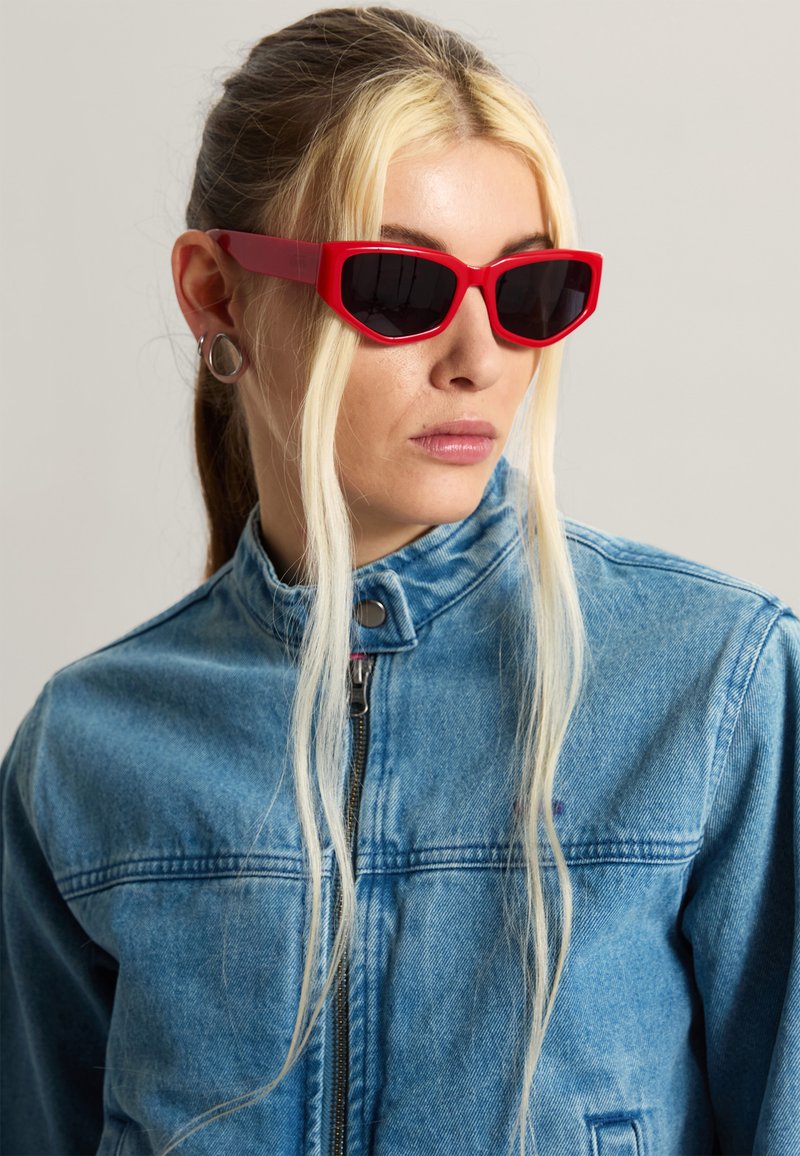 Mujer con cabello rubio largo que lleva gafas de sol rojas angulares, pendientes plateados y una chaqueta azul de mezclilla con cremallera, mirando hacia un lado.