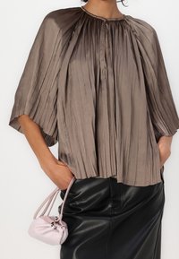 Femme portant un chemisier plissé taupe à manches à longueur de coude et une jupe en cuir noire, tenant un petit sac à cordon rose.