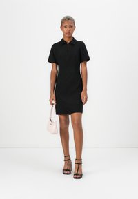 Black polo dress with short sleeves and a front zipper. Worn with black strappy heels and holding a small pink handbag.