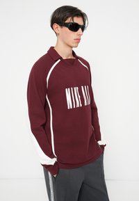 Burgundska polo majica s dolgimi rokavi z belim logotipom. Ima bele stranske črte in ovratnik. Model nosi črne sončne očala in siva hlače.