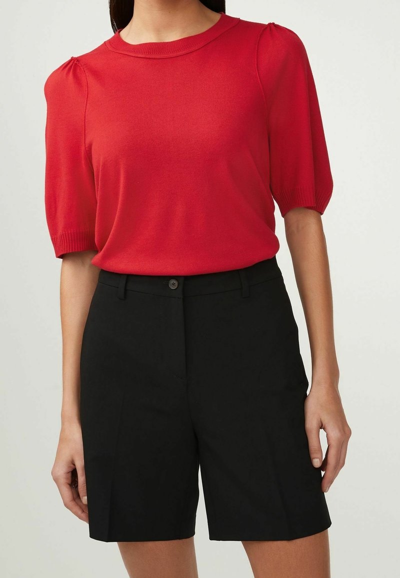 Haut en tricot rouge à manches courtes avec un col rond et des détails froncés aux épaules, associé à un short noir ajusté avec une fermeture à bouton à l'avant.