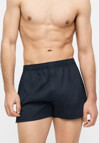 Άνδρας χωρίς πουκάμισο που φοράει μαύρα μποξεράκια με ελαστική μέση, στέκεται με τα χέρια χαλαρά μπροστά σε απλό φόντο.