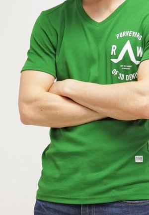 Mężczyzna w zielonej koszulce z białym napisem i niebieskich dżinsach, z założonymi na piersi skrzyżowanymi ramionami.