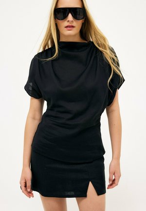 Mujer rubia con grandes gafas de sol negras, una blusa negra holgada y una falda corta negra con una abertura frontal, de pie sobre un fondo blanco.