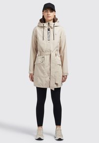 Beige monitoimijakku, jossa on kiristettävä huppu, vyö, kaksi etutaskua ja logo. Yhdistetty mustiin legginseihin ja vaaleisiin sneaker-kengiin.