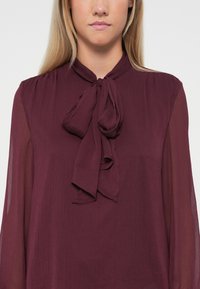 Blusa bordeaux con cravatta, realizzata in un tessuto leggero e testurizzato, con maniche lunghe trasparenti e un corpetto plissettato.