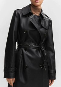 Schwarzer Kunstleder-Trenchcoat mit doppelt genähten Knöpfen, einem taillierten Gürtel und einem klassischen Kragen. Glatte Textur mit silberfarbenen Akzenten.
