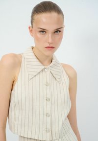 Korte mouwloze top van lichtbeige stof met fijne verticale strepen, voorzien van een brede kraag en een knoopsluiting aan de voorkant met lichte knopen.