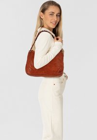 Borsa a tracolla in pelle marrone con una texture morbida e una forma curva, dotata di una tracolla intrecciata e dettagli metallici. Indossata sulla spalla.