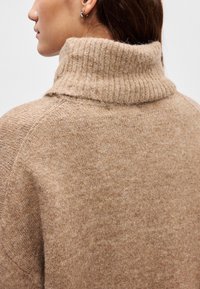 Bēšs adīts džemperis ar ribu raksta augstu apkakli. Mīksta tekstūra un nedaudz brīvs piegrieziens. Ar raglāna piedurknēm un minimālu detaļu daudzumu.