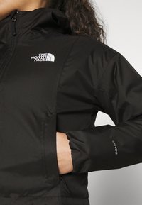 Zwarte jack met een ritssluiting aan de voorkant, met een logo op de linkerborst en 'Dryvent'-detail op de mouw. Het materiaal heeft een gladde textuur.