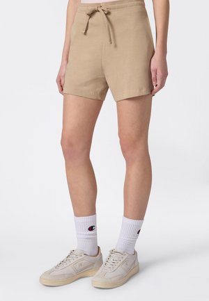 Persona che indossa pantaloncini beige con coulisse, calzini bianchi a metà polpaccio con logo e sneaker color avorio su sfondo bianco semplice.