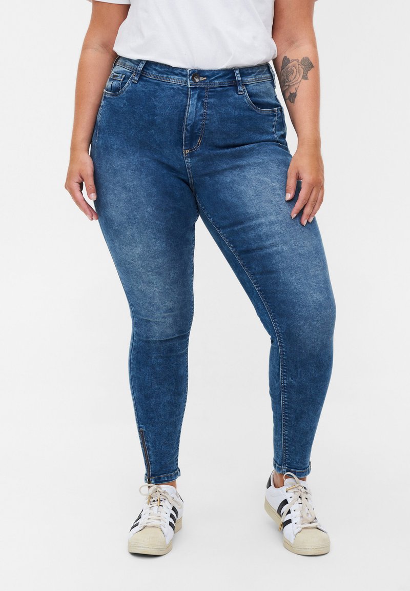 Vaqueros ajustados de color azul oscuro con cintura alta, que cuentan con bolsillos frontales y detalles con cremallera en los laterales de los tobillos, fabricados en un material de mezclilla.