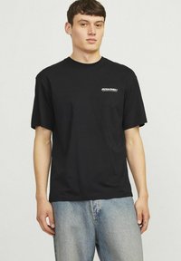 Sort bomuld t-shirt med korte ærmer, rund halsudskæring og et hvidt logo på venstre bryst. Matchet med lyseblå denim jeans.