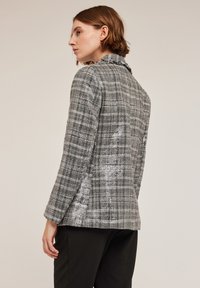 Blazer a quadri grigi e bianchi con un tessuto leggermente strutturato, dotato di colletto a sottocolla e chiusura con un bottone, mostrato da dietro.