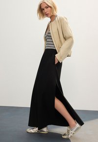 Bež bomber jakna preko crno-bijele prugaste majice bez rukava, uparena s crnom maksi suknjom sa bočnim prorezima i srebrnim tenisicama.