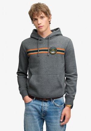 Jeune homme portant un sweat à capuche gris avec des rayures horizontales orange et vertes et un jean bleu, debout avec une main dans la poche.