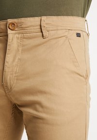 Pantalon en coton beige à coupe droite, avec une fermeture à bouton en bois, des détails cousus et une petite étiquette logo sur la ceinture.