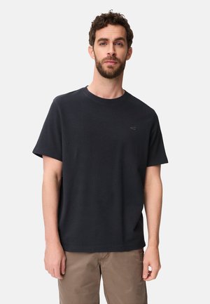 Man met kort krullend haar en baard, gekleed in een zwart T-shirt met korte mouwen en lichtbruine broek, staand tegen een effen achtergrond.