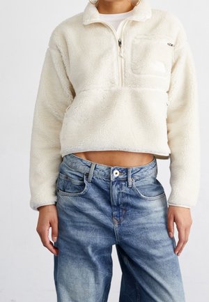 Osoba ubrana w białą polarową kurtkę zapinaną na zamek i luźne niebieskie dżinsy, kadrowane tak, aby pokazać odsłonięty brzuch i ręce spoczywające wzdłuż ciała.